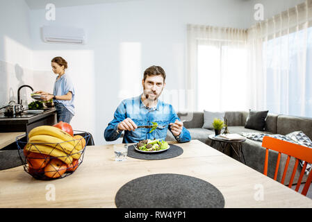 Man gesund essen Salat im Wohnzimmer der modernes Apartment mit Frau Geschirr auf dem Hintergrund Stockfoto