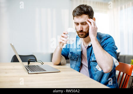 Mann trinken Arzneimittel aus der Kopfschmerzen während der Arbeit mit dem Laptop zu Hause Stockfoto