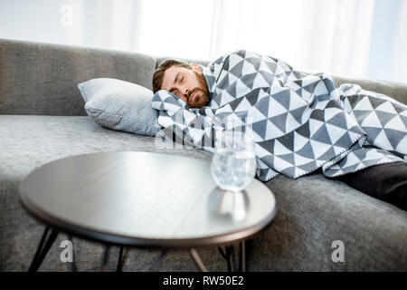 Mann krank auf der Couch liegen mit Decke mit Pillen und ein Glas Wasser auf dem Tisch zu Hause abgedeckt Stockfoto