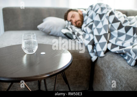 Mann krank auf der Couch liegen mit Decke mit Pillen und ein Glas Wasser auf dem Tisch zu Hause abgedeckt Stockfoto