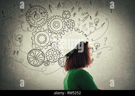 Harte denken nachdenkliche Frau Ingenieur suchen nachdenklich durch das Tragen von schützender helm und Zahnrad Zahnrad Gehirn Pfeile und Verwirrung als Gedanken über graue Wand. Stockfoto