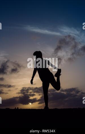 Zurück Blick auf die Silhouette der jungen Dame stretching Beine in Sportkleidung und wunderbaren Himmel mit Wolken in Abend Stockfoto