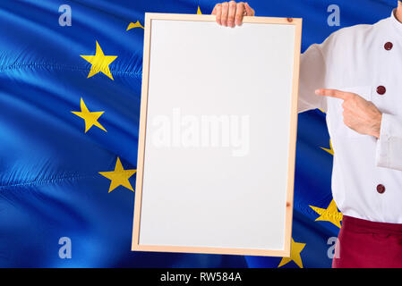 Koch Holding leeres Whiteboard Menü auf der Fahne der Europäischen Union Hintergrund. Cook tragen einheitliche zeigt Platz für Text. Stockfoto