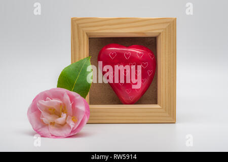 Platz Holzrahmen mockup mit einem rosa Blume mit einem grünen Blatt, die sich in einer Ecke des Bilderrahmens. Rotes Herz aus Stein im Inneren des Rahmens. Stockfoto