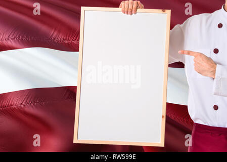 Lettland Koch Holding leeres Whiteboard Menü auf lettische Flagge Hintergrund. Cook tragen einheitliche zeigt Platz für Text. Stockfoto