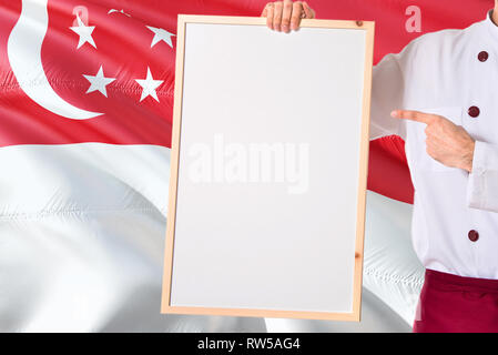 Singaporean Koch Holding leeres Whiteboard Menü auf Singapur Flagge Hintergrund. Cook tragen einheitliche zeigt Platz für Text. Stockfoto