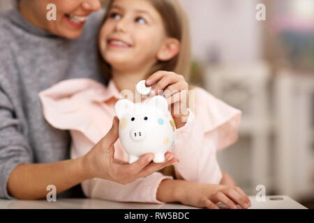 Junges Mädchen und ihre Mutter mit sparschwein am Tisch sitzen Stockfoto