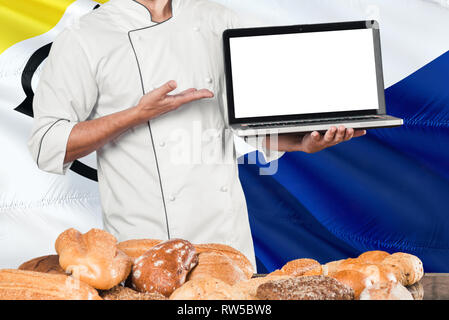Baker holding Laptop auf Bonaire Flagge und Brot Hintergrund. Koch tragen einheitliche zeigt leeren Bildschirm zu kopieren. Stockfoto
