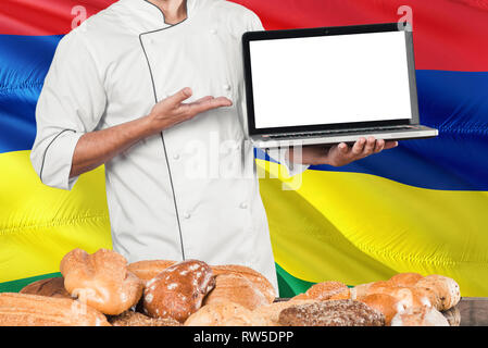 Mauritius Baker holding Laptop auf Mauritius Flagge und Brot Hintergrund. Koch tragen einheitliche zeigt leeren Bildschirm zu kopieren. Stockfoto