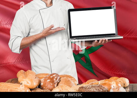 Marokkanische Baker holding Laptop auf Marokko Flagge und Brot Hintergrund. Koch tragen einheitliche zeigt leeren Bildschirm zu kopieren. Stockfoto