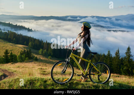 Junge weibliche Reiterin stehend mit gelben Fahrrad in die Berge, das Tragen von Helm, mit Blick auf das Tal auf Sommermorgen. Nebligen Berge, Wälder auf den verschwommenen Hintergrund. lifestyle Konzept Stockfoto