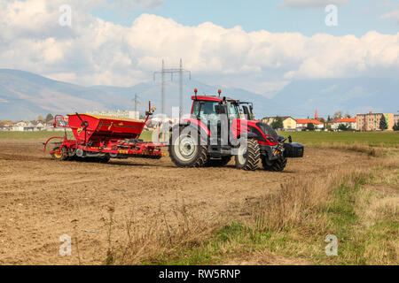 Red Tractor Aussaat, ziehen seeder Anhänger hinter, auf trockenem, einige Häuser und Berge im Hintergrund. Stockfoto