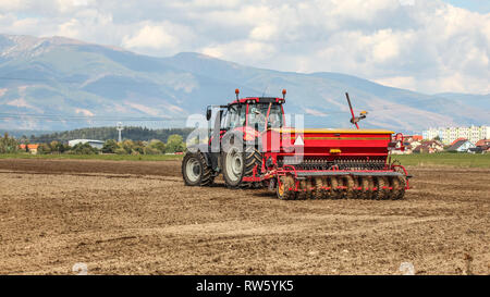 Red Tractor Aussaat Anhänger hinter ziehen über einem leeren Feld. Einige Dorf und Berge im Hintergrund. Stockfoto