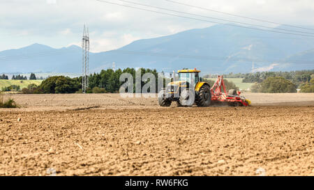 Gelbe Schlepper zieht Rot Aussaat über trockenes Feld schleppte, kleine Bäume und Berge im Hintergrund. Stockfoto
