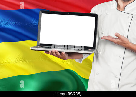 Mauritius Küchenchef holding Laptop mit leeren Bildschirm auf Mauritius Flagge Hintergrund. Cook Uniform tragen und zeigen Laptop für Kopieren. Stockfoto