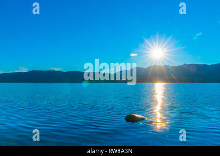 Sonnenaufgänge über ferne Murchisan Berge über den Lake Te Anau senden ein funkelndes Licht Spur über Wasser zu Lone rock South Island, Neuseeland Stockfoto