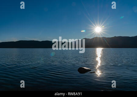 Sonnenaufgänge über ferne Murchisan Berge über den Lake Te Anau senden ein funkelndes Licht Spur über Wasser zu Lone rock South Island, Neuseeland Stockfoto