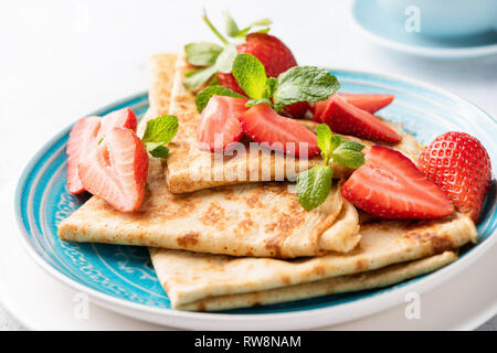 Blini oder Crepes mit Erdbeeren auf einem blauen Platte, Detailansicht. Leckere dünne Pfannkuchen zum Frühstück oder Dessert mit frischen roten Beeren serviert. Stockfoto