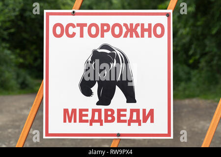 Warnschild auf Zaun in Russisch: Achtung Bären und schwarzem Piktogramm Kamtschatka Braunbär. Auf dem hintergrund der grünen Wald und Land Road Sign. Stockfoto