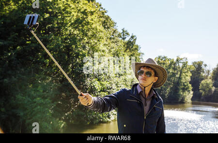 Junger Mann in Cowboy Hut und Sonnenbrille, selfie mit einem selfie Stick im Park auf sonnigen Sommertag Stockfoto