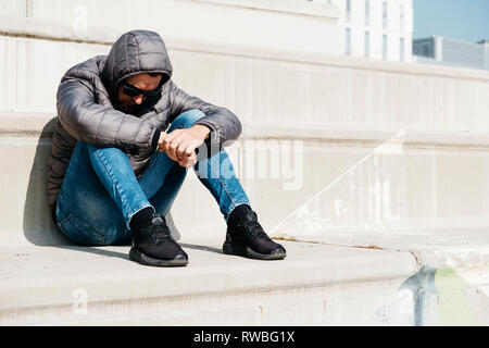 Ein junger weißer Mann, mit einer Jeans und einem Grau Hooded Jacket, gekräuselt oben sitzen auf einem im Treppenhaus Stockfoto