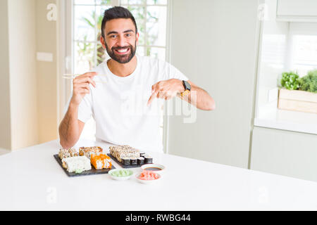Stattliche Hispanic Mann essen asiatische Sushi mit Stäbchen sehr glücklich zeigte mit Hand und Finger Stockfoto
