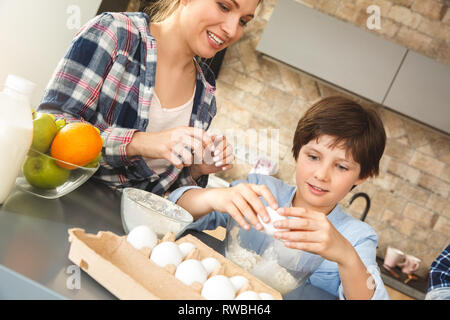 Mutter, Vater und Sohn zu Hause in der Nähe von Tisch in der Küche zusammen Mama und junge Aufschlagen der Eier in die Schüssel Lachen heiter close-up Stockfoto