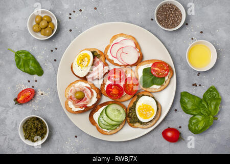 Varianten von Sandwiches mit Frischkäse, Pesto, Tomaten, Gurken, Ei, Speck, Rettiche auf grauem Hintergrund. Ansicht von oben Stockfoto