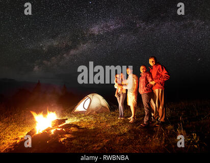 Nacht Camping in den Bergen. Touristen vor der brennenden Scheiterhaufen stehen. Beleuchtetes Zelt und Sternenhimmel für den Hintergrund. Frau mit in die Arme kleine Tochter. Tourismus und Freizeit Konzept. Stockfoto