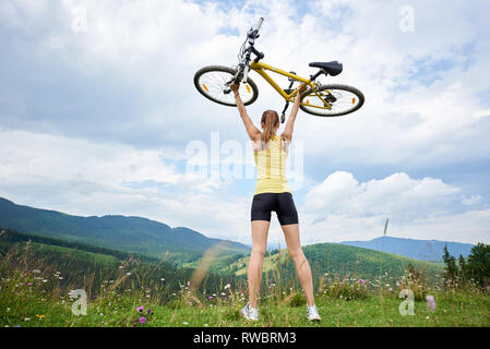 Rückansicht des Athleten Frau biker Holding gelben Mountainbike über einen Kopf, stehend auf einem grasbewachsenen Hügel, genießen Sommertag in den Bergen. Outdoor Sport, Lifestyle Konzept Stockfoto