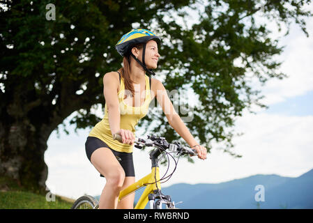 Attraktive Frau Radfahrer reiten auf gelben Mountainbike unter Big Tree, das Tragen von Helm, genießen Sommertag in den Bergen. Outdoor Sport, Lifestyle Konzept Stockfoto