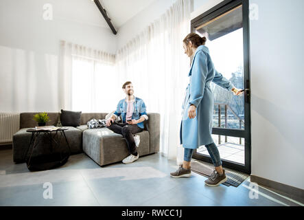 Junge Frau im blauen Mantel nach Hause kommen und ihr Mann auf der Couch im Wohnzimmer Stockfoto