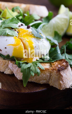 Vegetarisches Sandwich mit pochiertem Ei, Avocado, Rucola auf geröstetem Brot, gesundes Frühstück oder einen Snack auf rustikalen Holzbrett, selektiver Fokus, in der Nähe Stockfoto