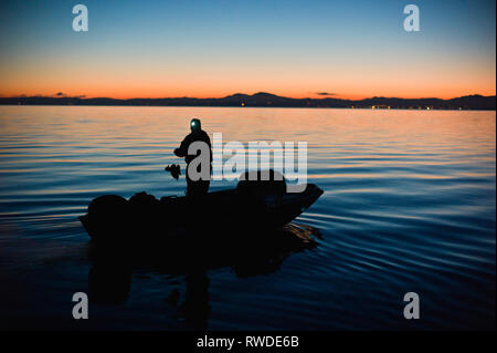 Mann mit einem Scheinwerfer steht in der Mitte ein Boot und sammelt in entelockvögel beim Schwimmen in einem See, wie die Sonne. Stockfoto