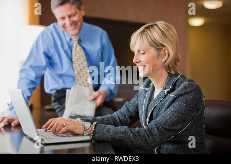 Geschäftsfrau am Schreibtisch und Typen auf einem Laptop sitzen, während die männlichen Kollegen sitzstangen am Rande der Schreibtisch in einem Hotel während einer Geschäftsreise. Stockfoto