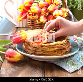 Pfannkuchen mit Kaviar und Butter in der Hand einer Frau, einen Strauß mit frischen Tulpen und eine hölzerne Hintergrund. Traditionelle russische Speisen für Karneval, Stockfoto