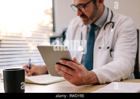 Reife männliche Arzt Holding eine digitale tablet Notizen in seinem Tagebuch während in seinem Büro zu sitzen. Arzt sitzt an seinem Schreibtisch mit Tablet Computer und Stockfoto