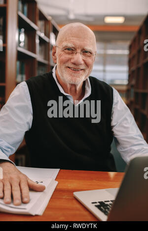 Lächelnd älterer Mann in der Bibliothek mit Buch und Laptop. Nahaufnahme eines älteren Mannes in einem Klassenzimmer und lernen sitzen. Stockfoto