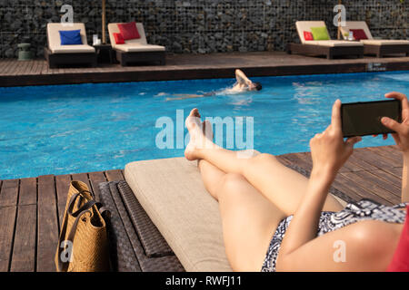 Schöne Dame mit Handy liegen auf Stuhl mit Mann Schwimmen im Pool. Stockfoto