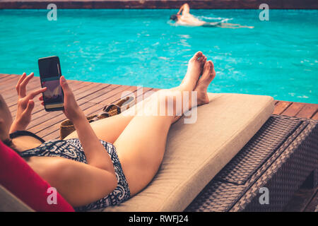 Schöne Dame mit Handy liegen auf Stuhl mit Mann Schwimmen im Pool. Stockfoto