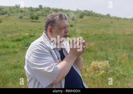 Schönes Portrait von Happy ukrainischen Landsmann Tabak Pfeife, während im Freien sitzen gegen hügeligen Frühling Weide Stockfoto