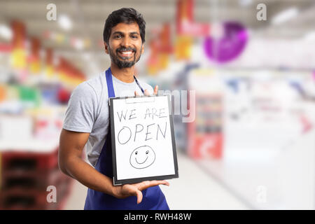 Freundlich indischen Supermarkt oder SB-Warenhaus Mitarbeiter präsentieren Zwischenablage mit öffnen wir sind Text und Smiley-face-Zeichnung Stockfoto