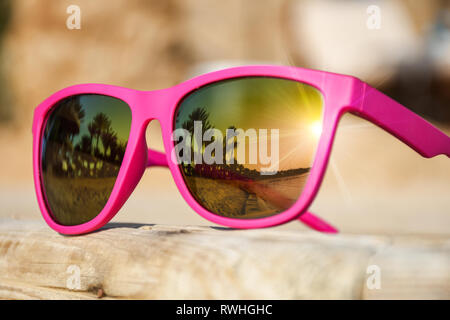 Moderne rosa Sonnenbrille auf einen Holzsteg mit schönen Reflexion der Strand mit Palmen und Sonnenuntergang Stockfoto