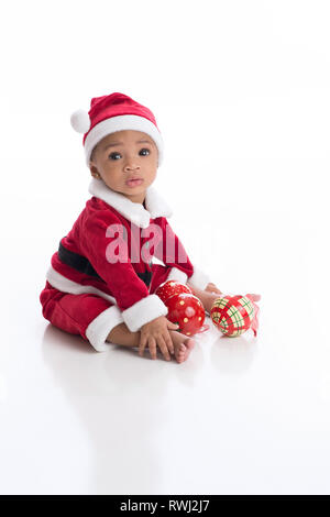 Sechs Monate alten Baby Mädchen mit einem Weihnachtsmann Kostüm. Sie sitzt auf einem weißen, nahtlose Hintergrund mit Weihnachtsschmuck. Stockfoto