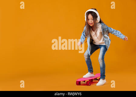Stilvolles kleines Kind Mädchen in Kopfhörer mit Skateboard in Jeans Kleidung auf orangefarbenem Hintergrund Stockfoto