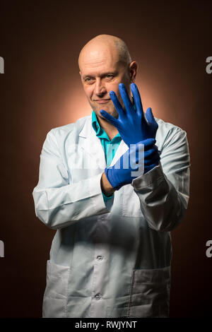 Arzt 50 s trägt einen weissen Mantel erlebt, trägt blaue Gummihandschuhe und lächelnd Stockfoto