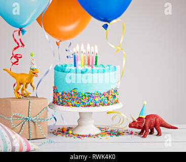 Childs Birthday Party Szene mit blauer Kuchen, Geschenkbox, Spielzeug Dinosaurier, Hüte und bunte Luftballons über Hellgrau. Stockfoto