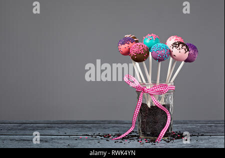 Sortiment an Kuchen Pop in ein Glas mit Schokolade besprüht und eine rosa Schleife über einen grauen Hintergrund. Stockfoto