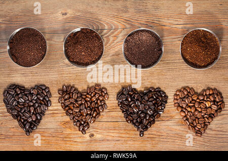 Ansicht von oben von vier Sorten von gerösteten Kaffeebohnen in der Form der Herzen mit ihren frisch gemahlenen Pendants in kleinen Schälchen auf braunem Holz- Stockfoto