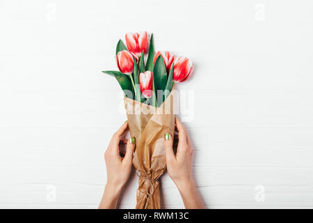Woman's Händen grüne Maniküre holding Bouquet von roten Tulpen Blumen in Braun Kraftpapier auf weißem Holz- Hintergrund, Ansicht von oben. Stockfoto